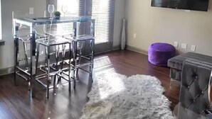 Een moderne woonkamer met een eethoek, een plafondventilator, een flatscreen-televisie en een groot raam met luxaflex.