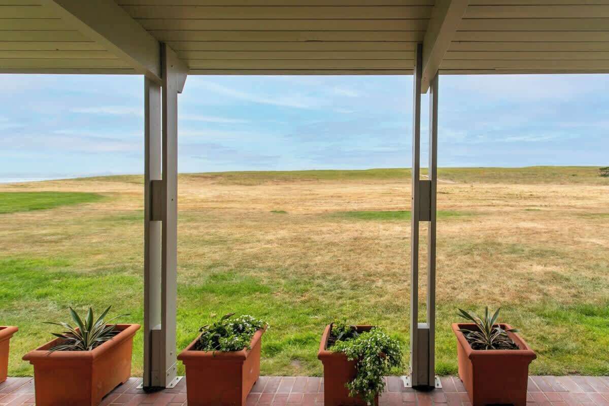 A covered outdoor area with a view of a grassy field, supported by columns, and potted plants.