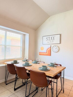 Ein Essbereich mit einem Holztisch, gedeckt für vier Personen, vier braunen Stühlen, einem Fenster mit Jalousien und Wandbildern, darunter eine Uhr und ein Schild mit der Aufschrift 'STAY mobile'.