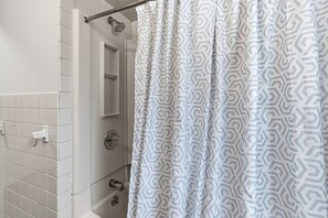 Un baño con paredes de azulejos blancos, una ducha con cortina de baño estampada en blanco y gris, y un perchero visible.