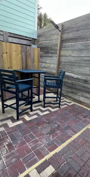 Une terrasse extérieure avec une table et deux chaises, une clÎture en bois et un sol en briques.
