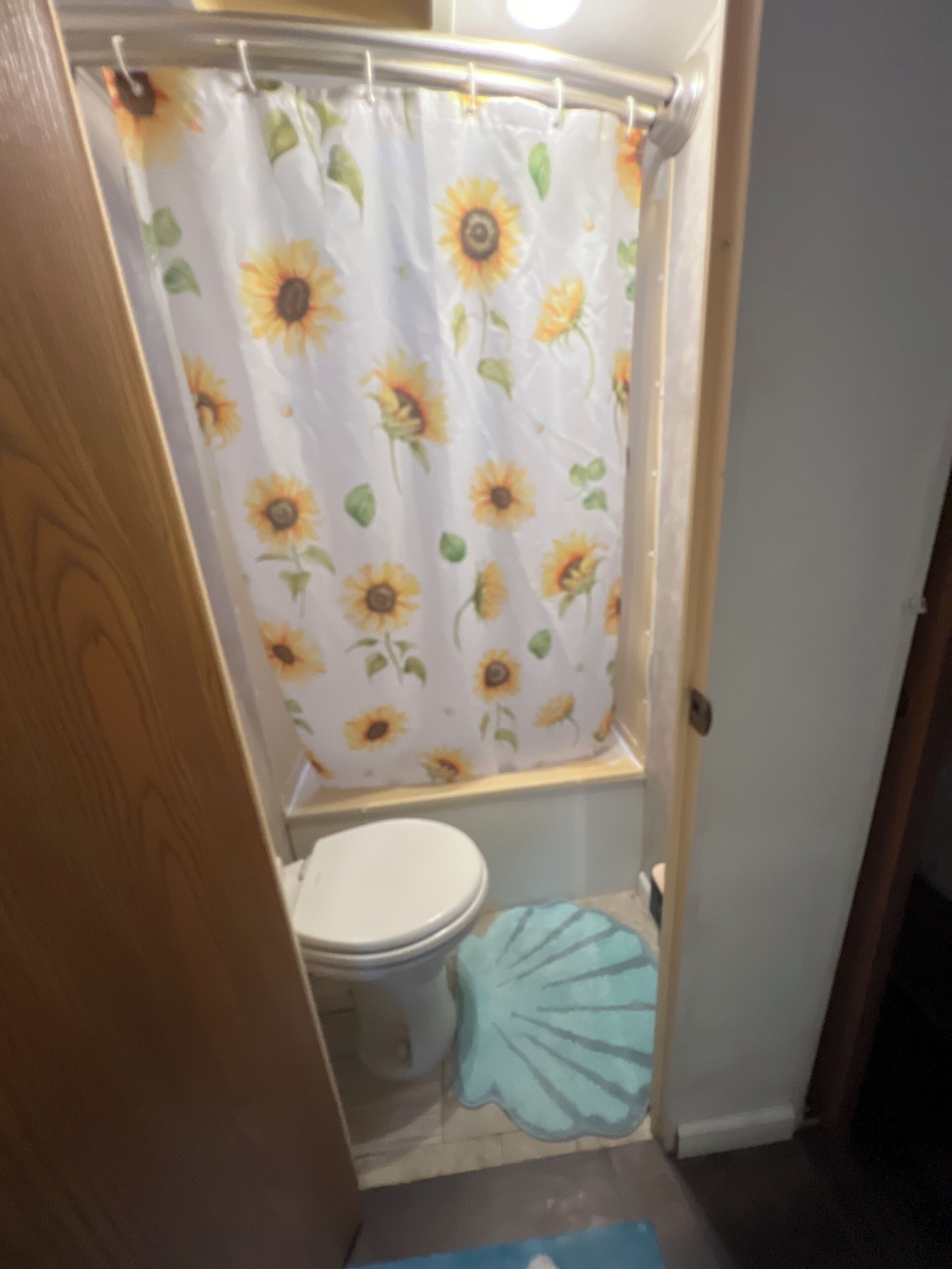 A bathroom with a toilet, a shower curtain with sunflowers, and a blue mat.