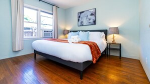 Quarto com cama, janela com cortinas, mesa de cabeceira com luminária e pintura abstrata pendurada na parede.