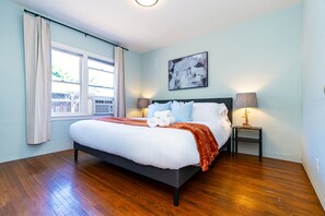 Un dormitorio con una cama, una ventana con cortinas, una mesita de noche con lámpara y un cuadro abstracto colgado en la pared.