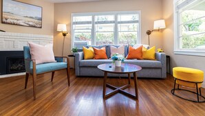 Uma sala de estar com um sofá, duas poltronas, uma mesa de centro e um banquinho amarelo. Há duas luminárias de chão, uma lareira e uma janela grande com vista para o verde.