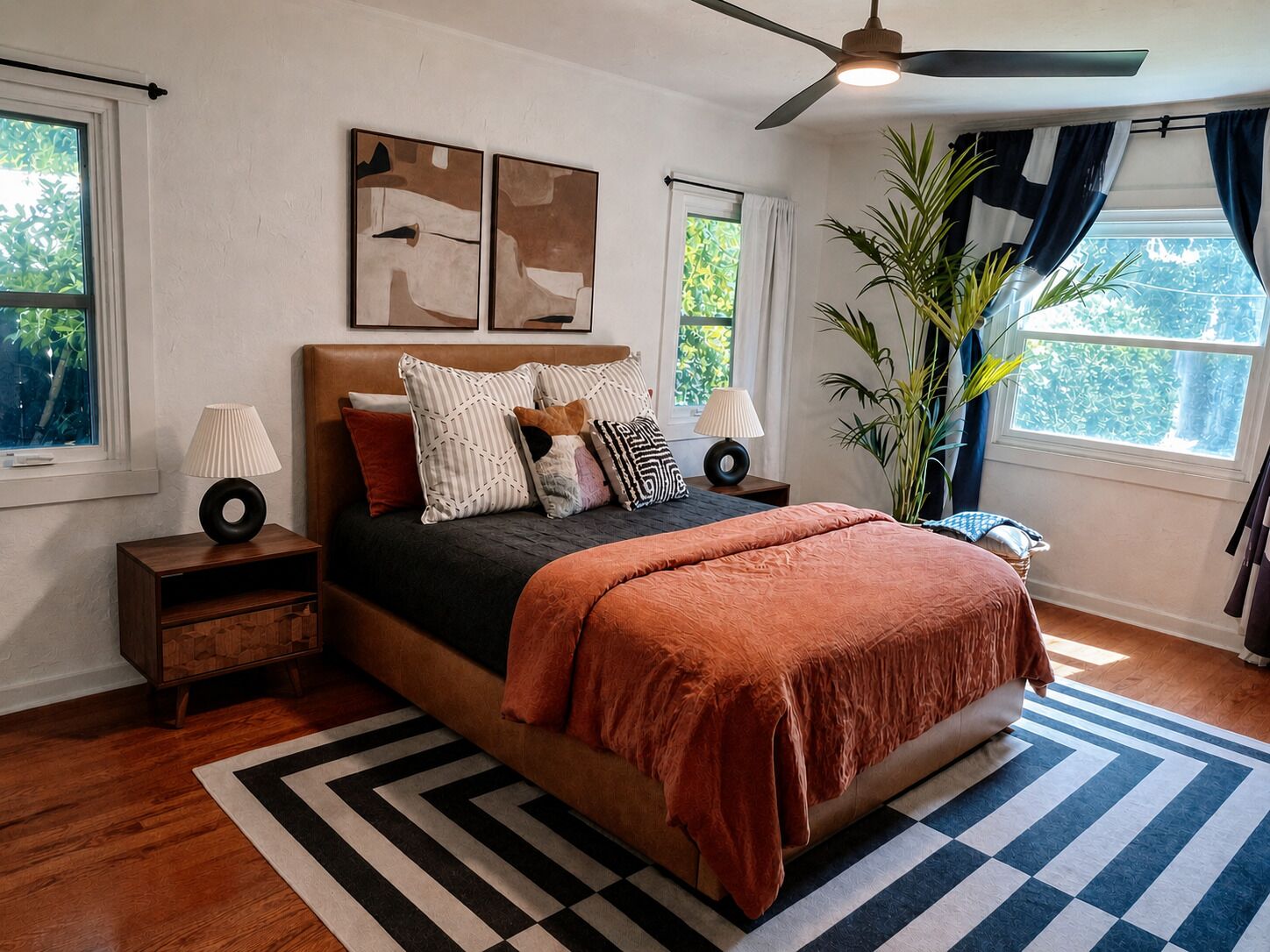 Un dormitorio con cama, mesitas de noche, ventilador de techo, obras de arte, una planta en maceta y vistas a zonas verdes al exterior.
