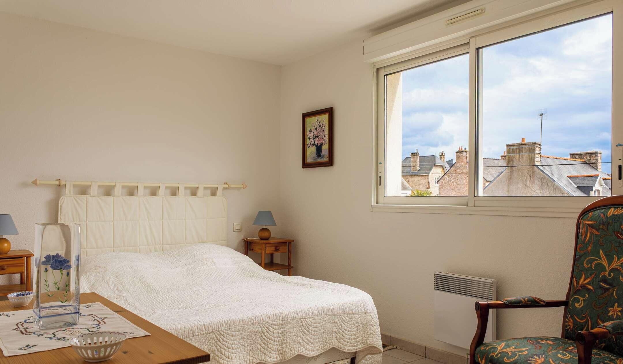 Ein Schlafzimmer mit einem Bett, Nachttischen, einem Sessel, einem Fenster mit Blick auf Gebäude und einer Heizung.