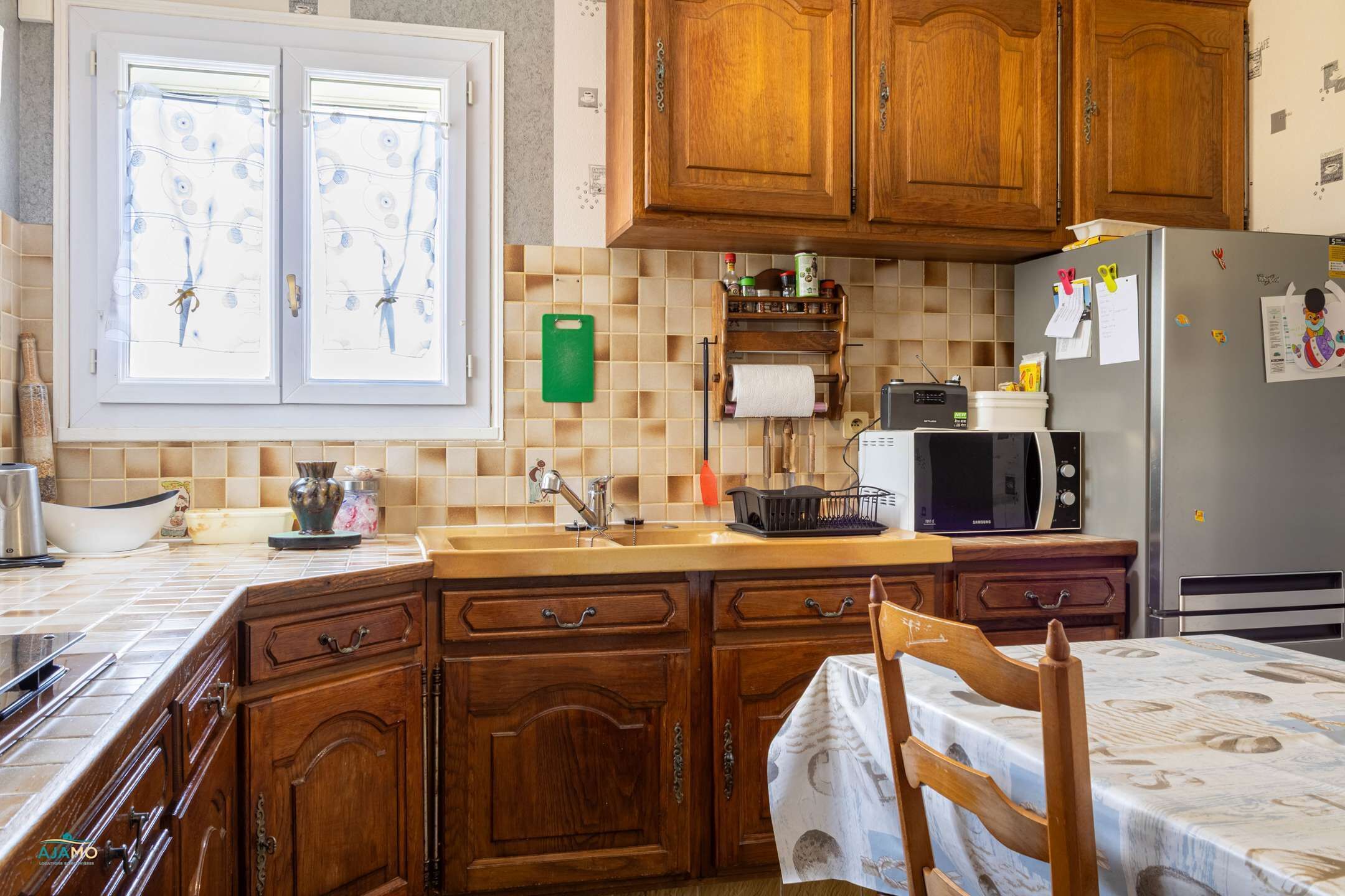 Une cuisine avec des meubles en bois, un four à micro-ondes, un réfrigérateur et une fenêtre avec des rideaux.