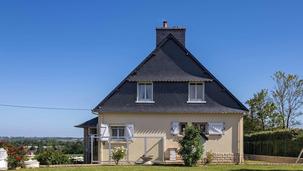 Une maison à deux étages, avec un toit sombre, des volets blancs et un petit jardin.