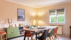 Un coin repas avec une table dressée pour quatre personnes, un meuble vert, un tableau encadré et une fenêtre avec des stores.