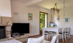 A living room with a stone fireplace, a flat-screen TV, a dining table with chairs, and artwork on the walls. - Cozy house for 9 people between beach and nature (FREHEL)