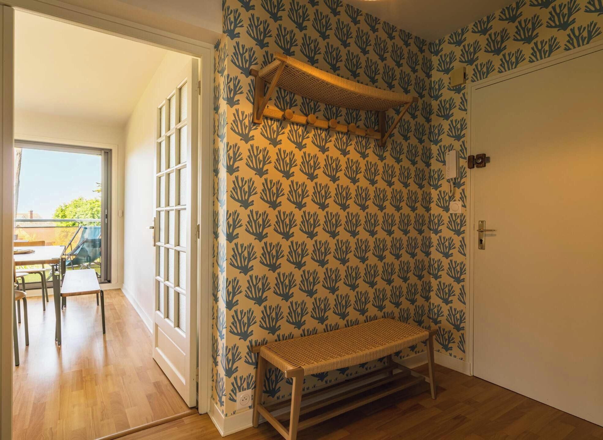 A room with a patterned wallpaper, a bench, a door, and a view of an outdoor area with a table and chairs.