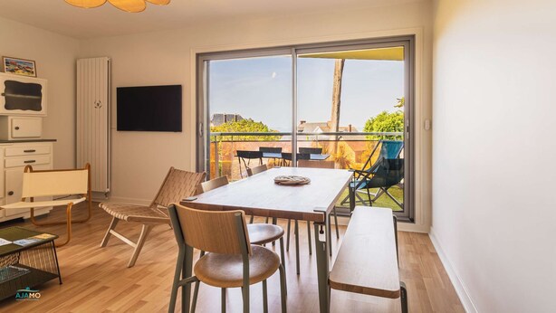 Ein modernes Wohnzimmer mit einer großen Glasschiebetür zum Balkon, einem Esstisch mit Stühlen, einer Couch und einem an der Wand befestigten Fernseher.