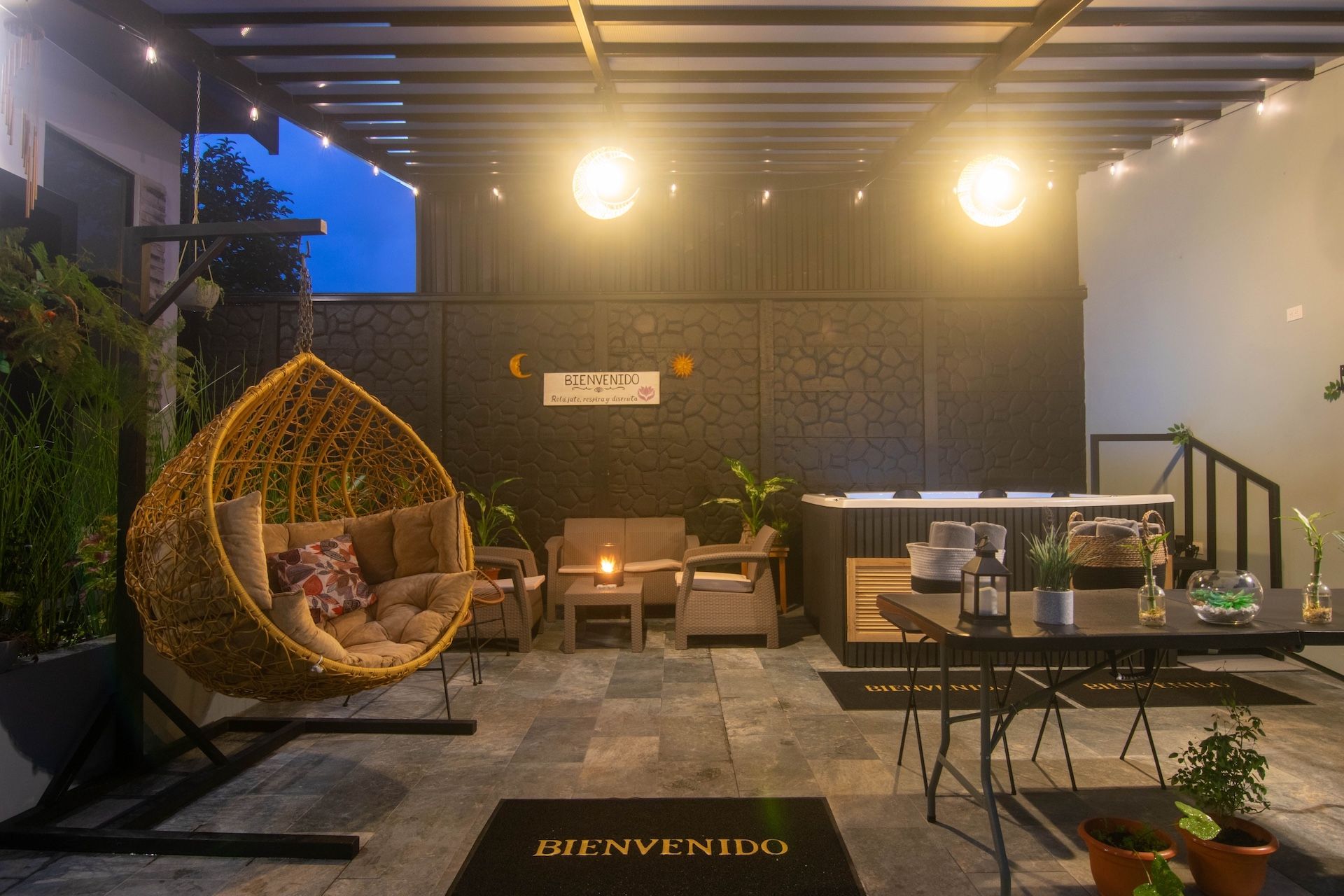 Un rincón acogedor al aire libre con una silla de mimbre, una mesa con cafetera y un letrero que dice 'BIENVENIDO'.