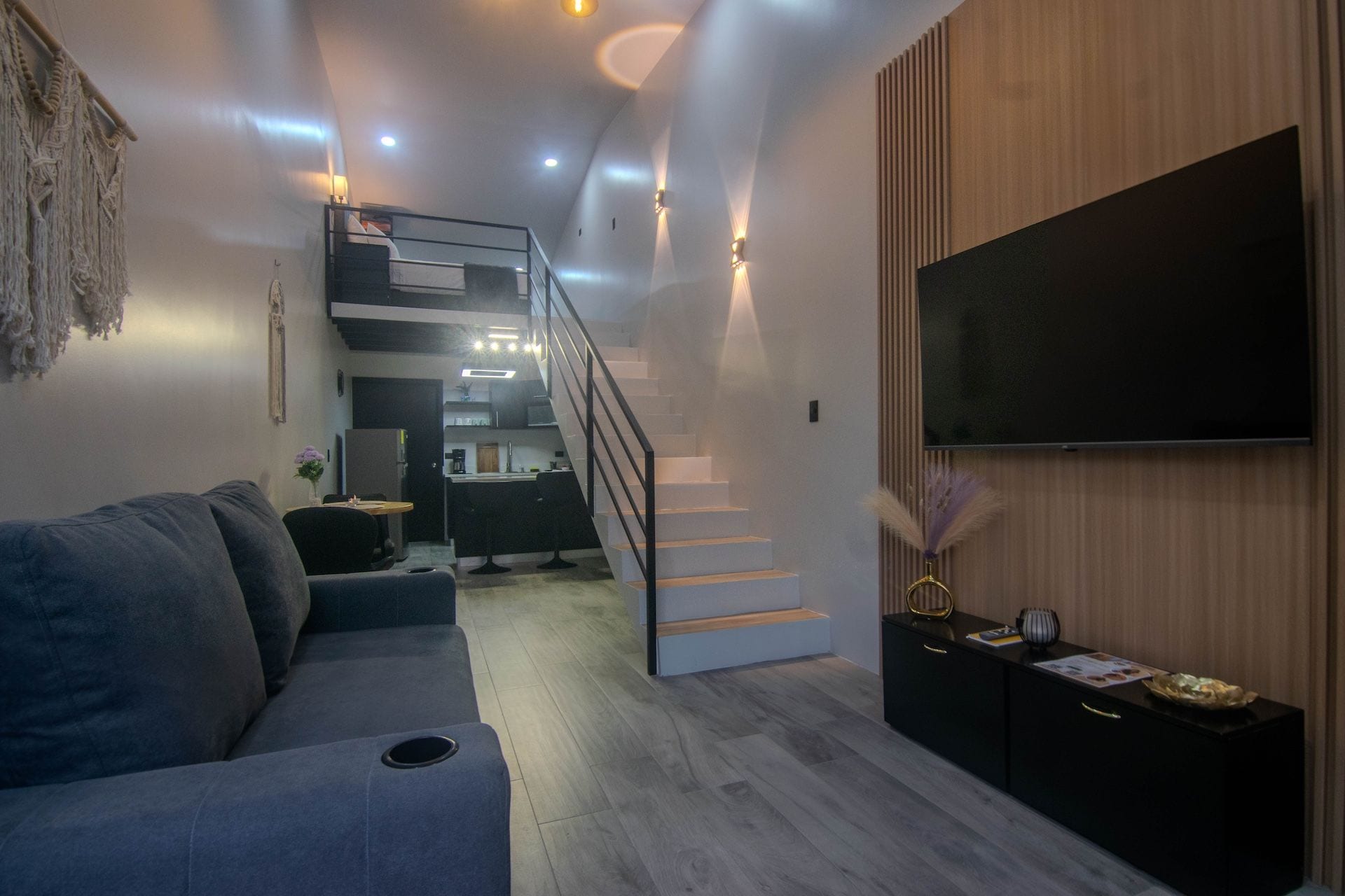 Una sala de estar moderna con un sofá, un televisor de pantalla plana, una escalera que lleva a una cocina y una pared de acento de madera.
