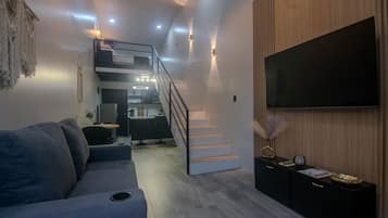 Una sala de estar moderna con un sofá, un televisor de pantalla plana, una escalera que lleva a una cocina y una pared de acento de madera.