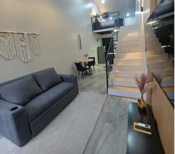 A modern living room with a grey sofa, a small dining table with chairs, a staircase, and a macrame wall hanging.