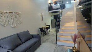 Una sala moderna con un sofá gris, una pequeña mesa de comedor con sillas, una escalera y un colgador de macramé en la pared.