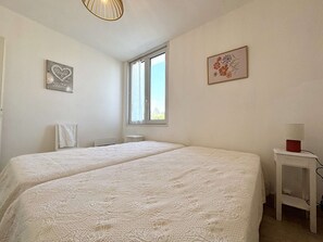 Ein Schlafzimmer mit zwei Betten, einem Fenster, einem Heizkörper, einem kleinen Tisch mit einer Lampe und Wandbildern.