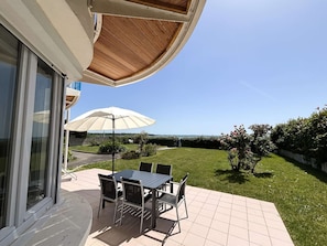 Eine Terrasse mit Tisch und Stühlen, einem weißen Sonnenschirm und Blick auf das Meer.
