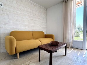 Ein Wohnzimmer mit einer Couch und einem Tisch, einem Fenster mit Vorhängen und Blick ins Grüne.