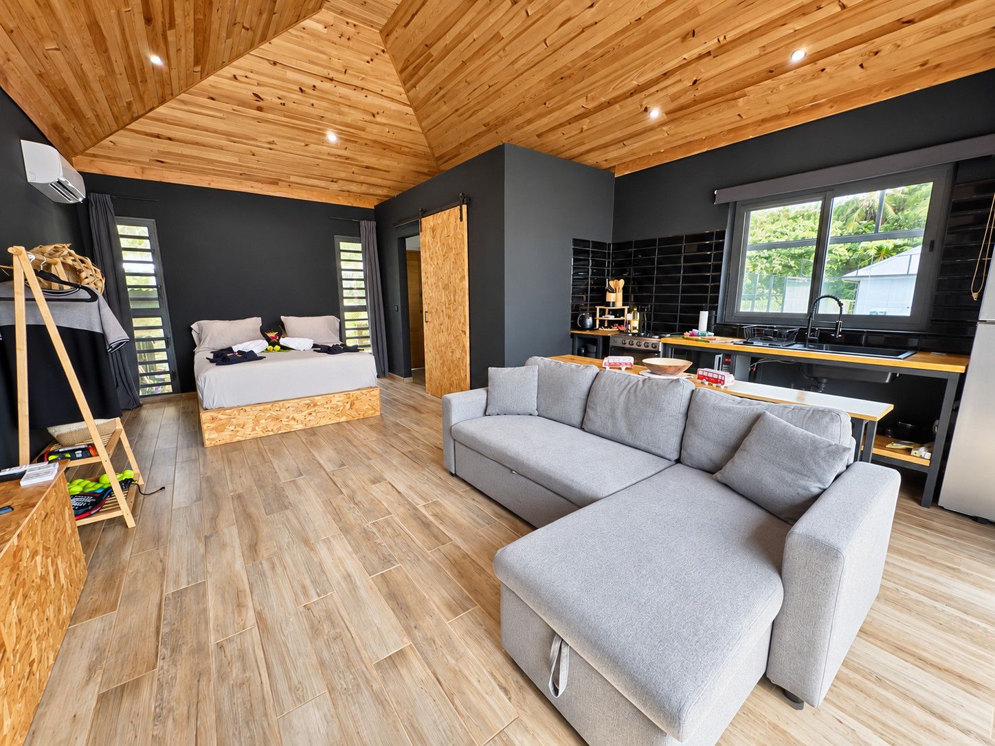 Un intérieur moderne avec un plafond en bois, un canapé gris, un lit et un coin cuisine.