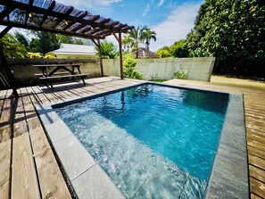 En rektangulær swimmingpool med klart, blåt vand, omgivet af en træterrasse og et pergola.