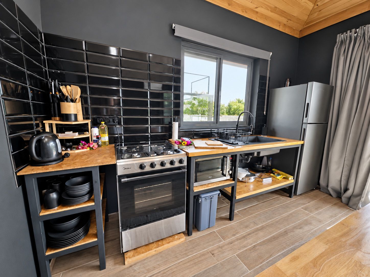 A modern kitchen with black tiled backsplash, stainless steel appliances, wooden countertops, and a window providing natural light.