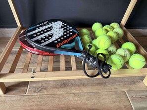 Ein Tennisracket mit blauem Griff und einem schwarz-roten Rahmen, liegend auf einem hölzernen Gestell mit einer Sammlung gelber Tennisbälle.