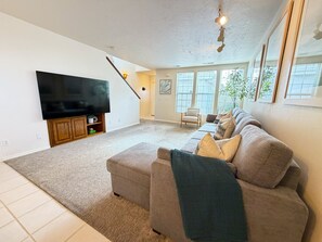 Ein Wohnzimmer mit einem großen Flachbildfernseher, einem Sofa, einer Treppe und großen Fenstern.