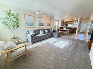 Ein Wohnzimmer mit einem grauen Ecksofa, einem Holzstuhl, einer Topfpflanze und einem Essbereich mit Tisch und Stühlen.