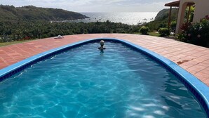 A swimming pool with clear blue water, surrounded by a tiled deck and lush greenery, overlooking a coastal landscape.