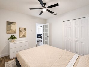 A bedroom with a bed, a ceiling fan, a dresser, a plant, and a door leading to another room. - Cozy 2BR Retreat w/Private Patio Near Downtown, Airport & Hospitals (Miami)
