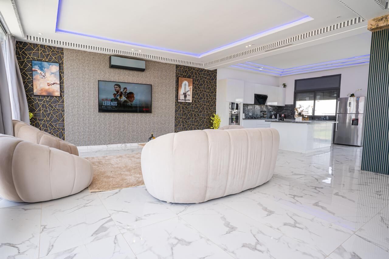 A modern living room with a flat-screen TV, comfortable seating, a kitchen area, and a marble floor.