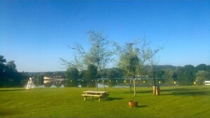 A grassy area with a picnic table, trees, a small body of water, and a distant building. - Mobile Home Lodge (Bonzée)