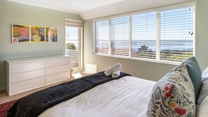 A bedroom with a bed, a dresser, a window with blinds, and a view of the sea. - Coastal Retreat Family Home | Sea Views + Parking (Christchurch)