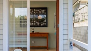 A wooden deck with a potted plant, a mat, and a door leading to an interior space with a bench and a wall-mounted picture. - Coastal Retreat Family Home | Sea Views + Parking (Christchurch)
