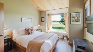 Een slaapkamer met een houten plafond, een bed met wit beddengoed, een nachtkastje, een televisie en een raam met gordijnen.