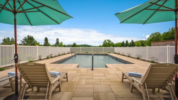 Une piscine avec deux chaises longues et deux parasols, entourée d’une clôture blanche et sous un ciel dégagé.