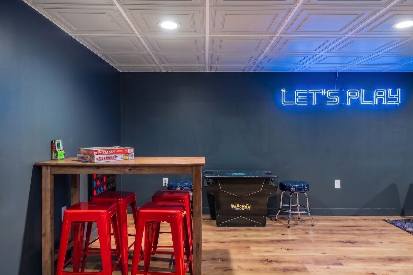 A room with a neon sign reading 'LET'S PLAY,' a wooden table with Monopoly and other board games, red bar stools, and a pool table.