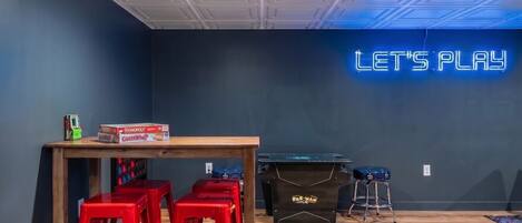 A room with a neon sign reading 'LET'S PLAY,' a wooden table with Monopoly and other board games, red bar stools, and a pool table.