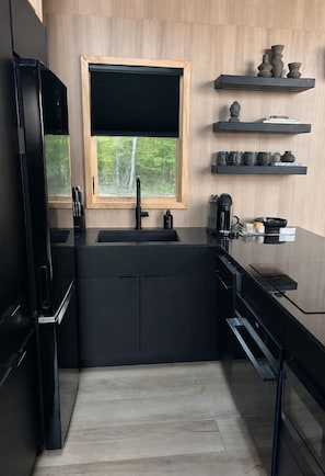 A modern kitchen with a black countertop, a window with a black blind, and wooden shelves above the sink. - Private Romantic Escape with Scenic Views + Fire Pit (Sparta)