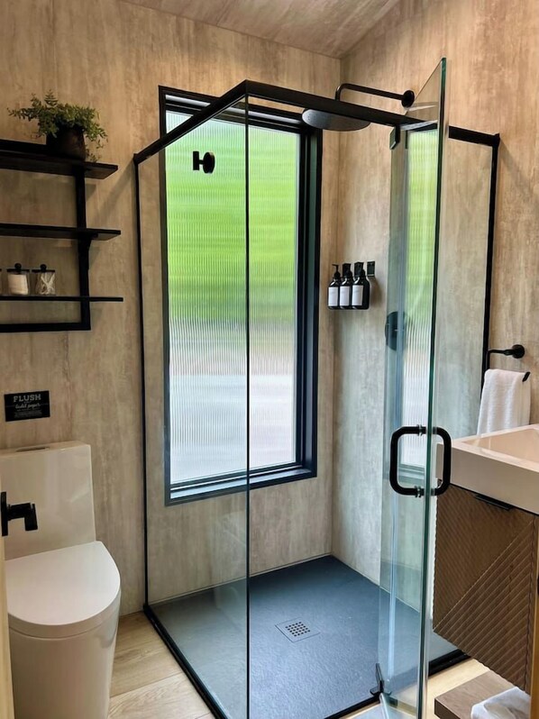 Un baño moderno con ducha envuelta en vidrio, inodoro, lavamanos y ventana con vistas a la vegetación.