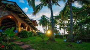 A brick building with arched walkways and a garden with palm trees and potted plants. - Peñiscola Villa - Beachfront Estate in El Zonte, Surf City - El Salvador (Chiltiupán)