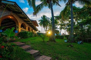 A brick building with arched walkways and a garden with palm trees and potted plants. - Peñiscola Villa - Beachfront Estate in El Zonte, Surf City - El Salvador (Chiltiupán)