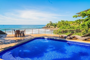 Piscina com vista para o oceano, uma mesa e cadeiras, e uma pérgula com plantas.