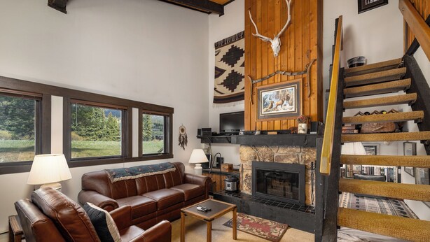 A living room with a fireplace, leather sofa, and a staircase. - Nordic Pines Retreat | Ski, Golf & Unwind (Big Sky)