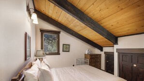 A bedroom with a wooden ceiling, a bed with white bedding, a nightstand, a lamp, a dresser, and a window with a view of trees. - Nordic Pines Retreat | Ski, Golf & Unwind (Big Sky)