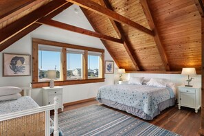 Quarto com teto inclinado, vigas de madeira, uma janela grande, cama com colcha estampada, mesas de cabeceira com luminárias e vista para o exterior.