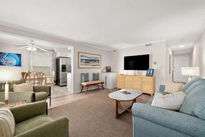 Ein Wohnzimmer mit einer Couch, einem Couchtisch, einem Fernseher, einem Essbereich mit Stühlen und einer Küche mit Kühlschrank.